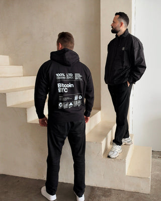 Schwarzer Bitcoin Label Hoodie von BITVESTIS in Rückansicht mit weißem Bitcoin-BTC-Print, daneben ein Model in schwarzer Bitcoin Freizeitjacke auf einer Treppe.