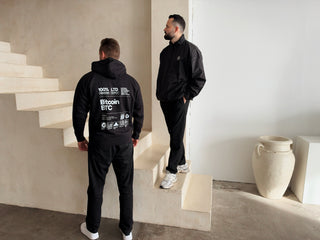 Schwarzer Bitcoin Label Hoodie von BITVESTIS in Rückansicht mit weißem Bitcoin-BTC-Print, daneben ein Model in schwarzer Bitcoin Freizeitjacke auf einer Treppe.