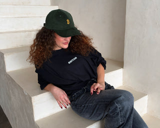 Dunkelgrüne Bitcoin Cord-Cap von BITVESTIS mit goldenem Bitcoin-Stick, getragen von einer Frau im schwarzen T-Shirt mit weißem Bitcoin-Brustprint.