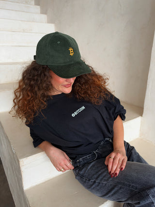 Dunkelgrüne Bitcoin Cord-Cap von BITVESTIS mit goldenem Bitcoin-Stick, getragen von einer Frau im schwarzen T-Shirt mit weißem Bitcoin-Brustprint.